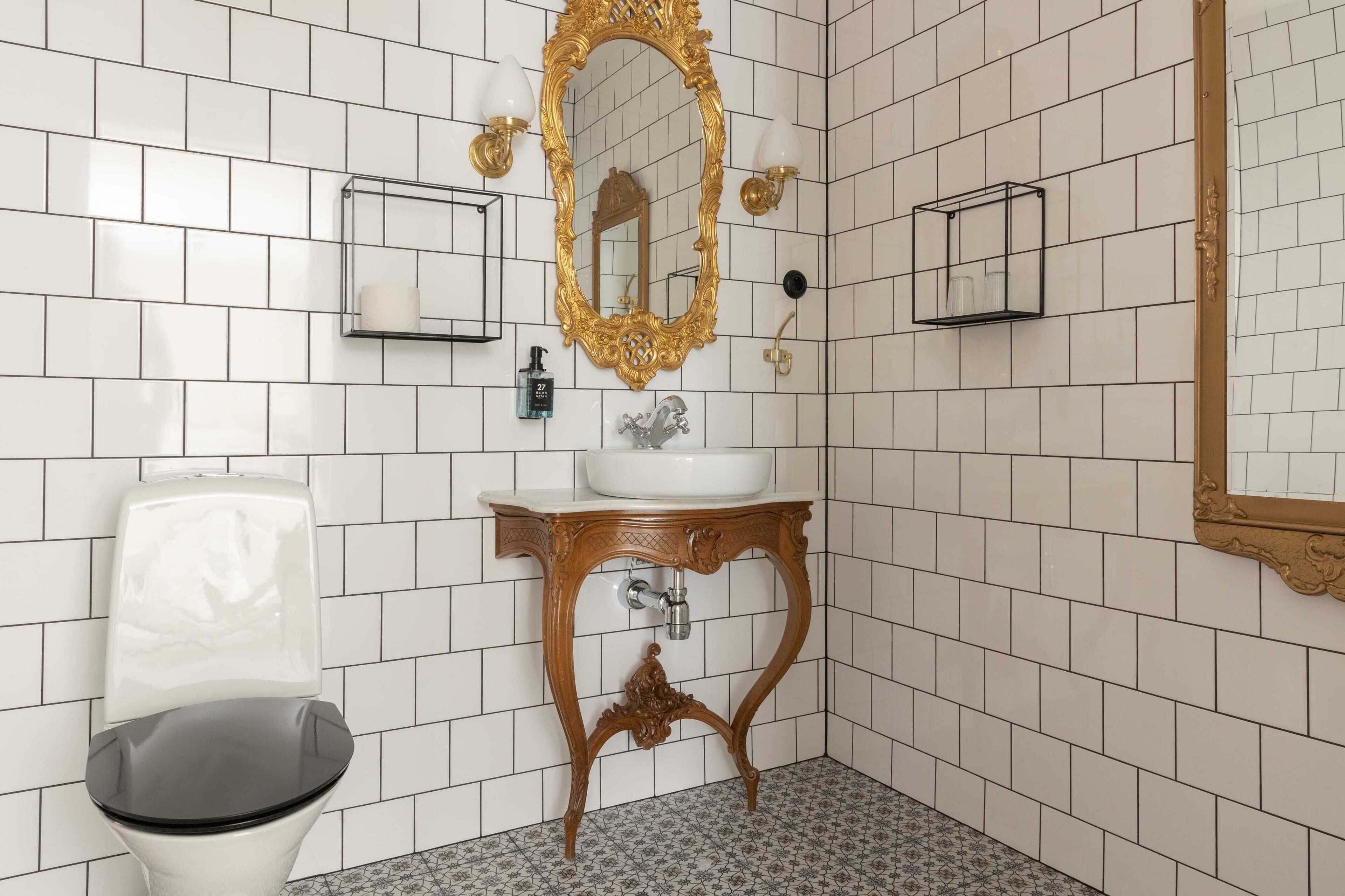 Un baño con paredes de azulejos blancos, un lavamanos blanco, un espejo con marco dorado y un inodoro negro.