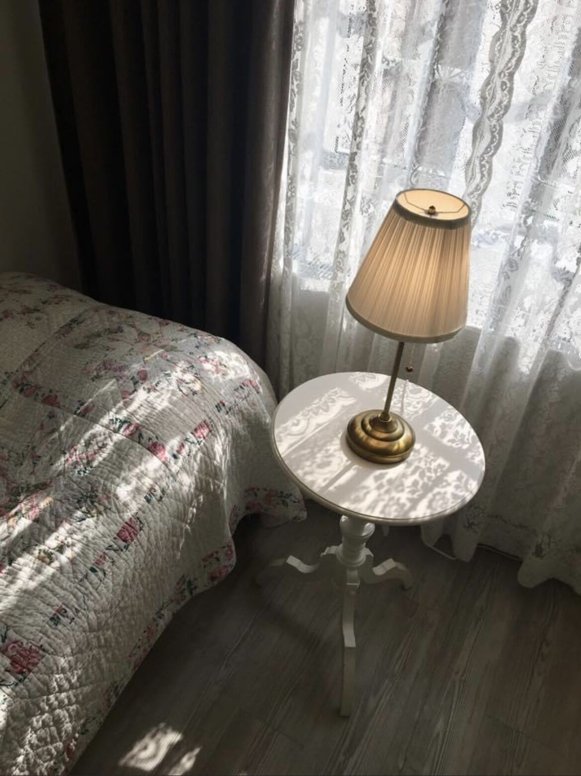 Una cama con una colcha estampada, una mesa redonda con una lámpara y cortinas de encaje.
