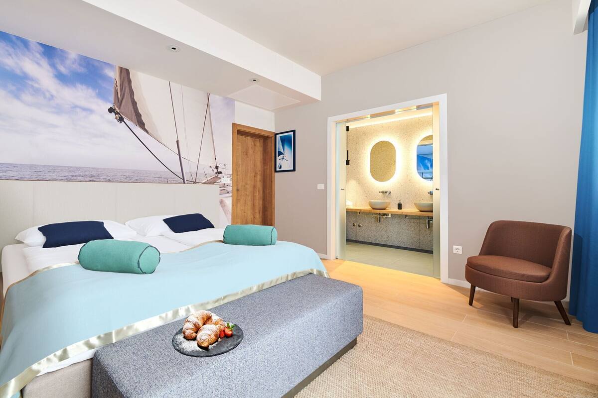 A modern bedroom with a bed, a bench with pastries, a chair, and a bathroom with a large mirror and sink.