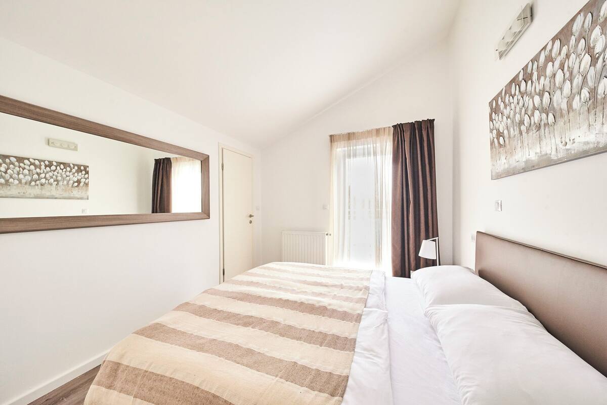A hotel room with a bed, a large mirror, a painting on the wall, and a window with curtains.