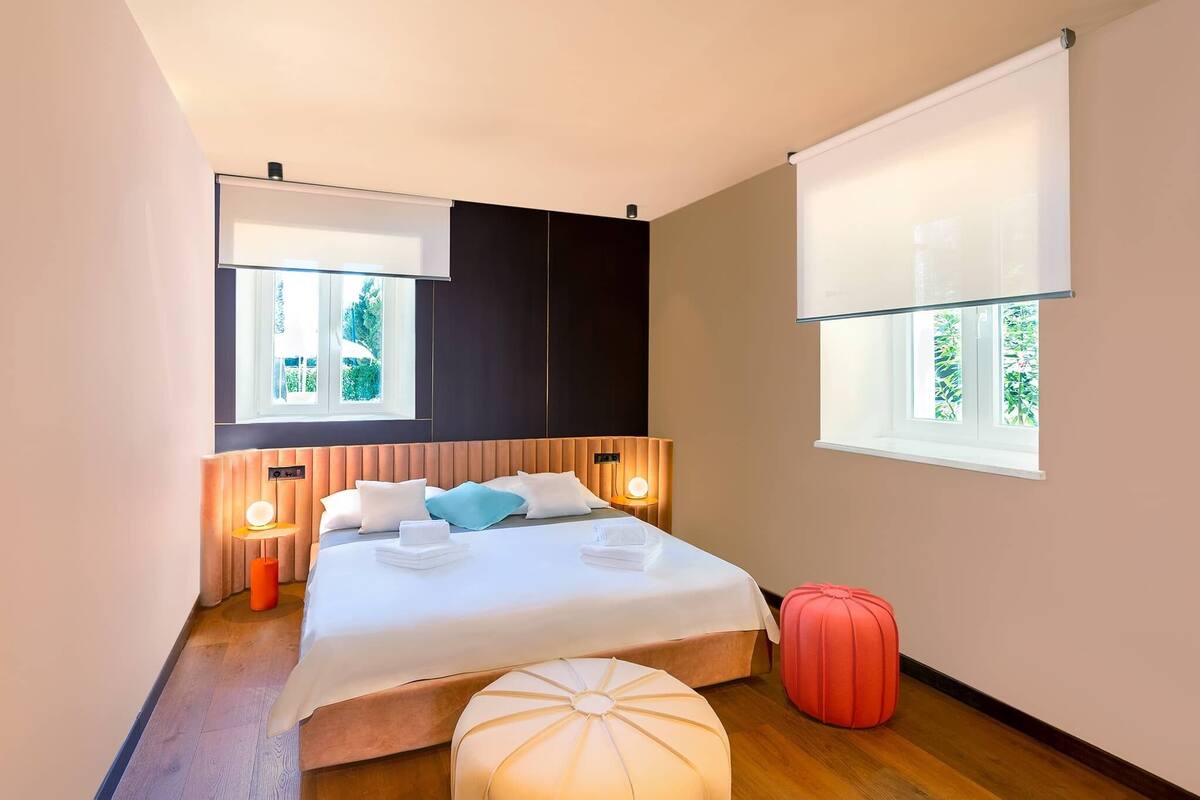A modern hotel room with a large bed, bedside lamps, a window with a view, and a red ottoman.