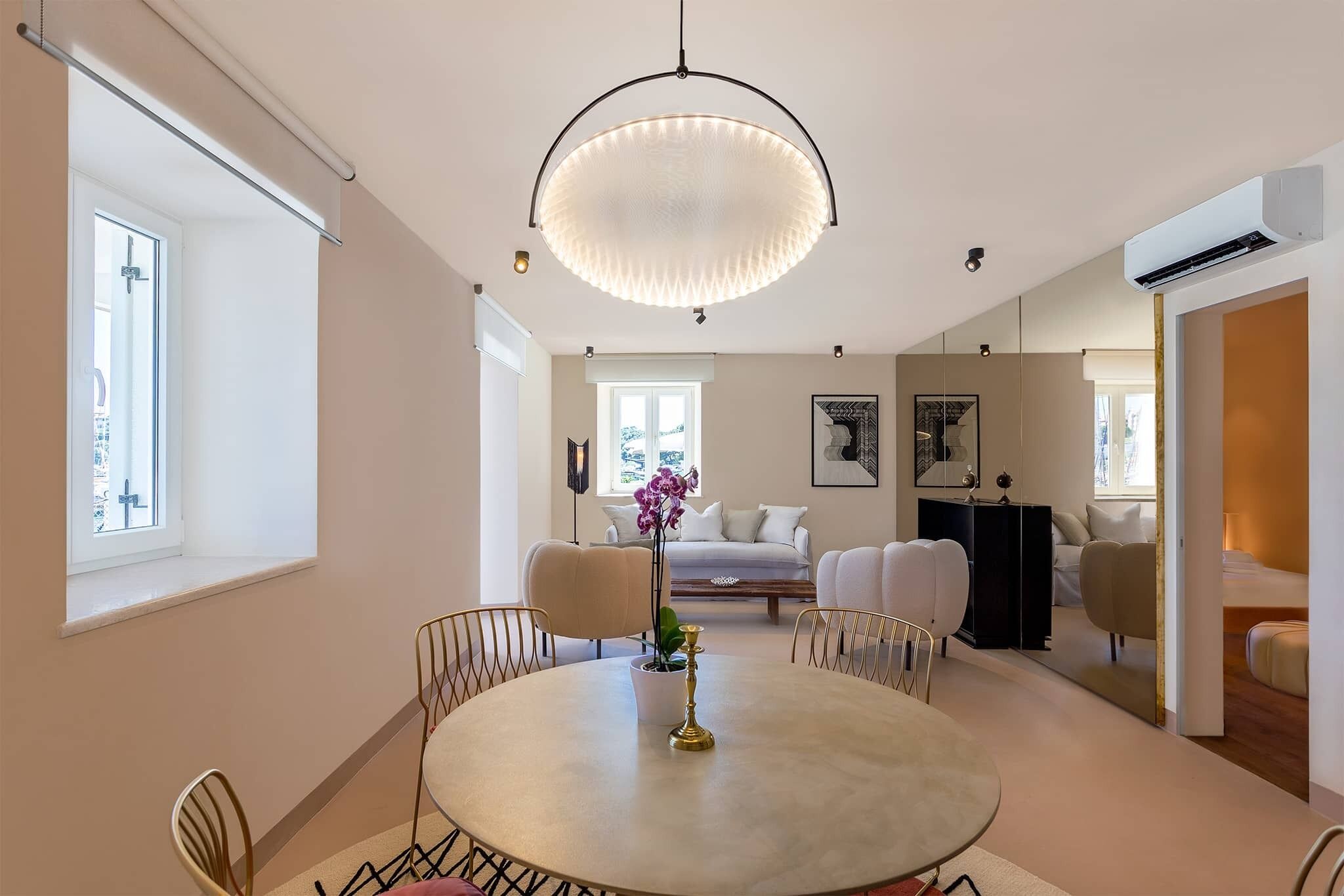 A modern living room with a round dining table, comfortable seating, a large chandelier, and a view of the city through a window.