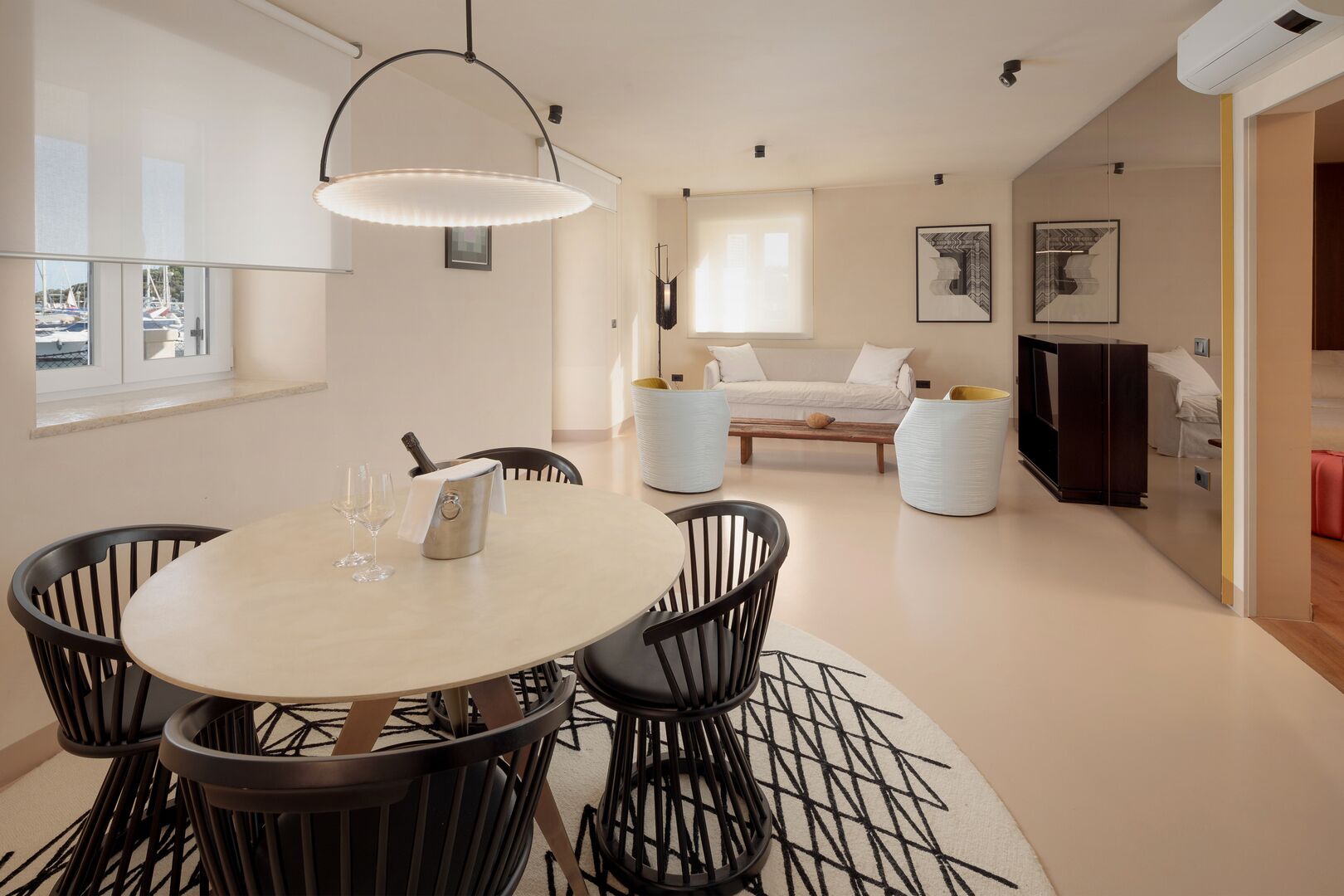A modern living room with a round dining table, black chairs, a white sofa, and a large window with a view of boats.