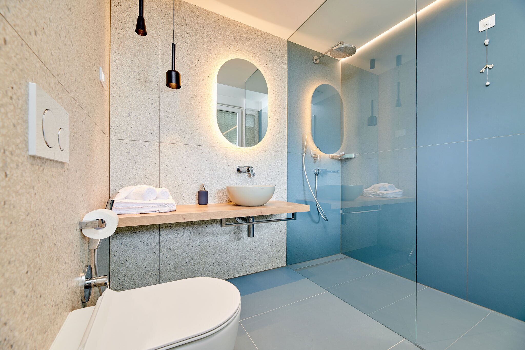 A modern bathroom with a freestanding toilet, a large oval mirror, a built-in sink with a vessel basin, and a glass-enclosed shower area.