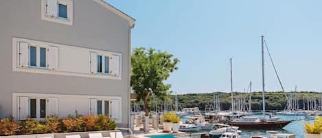 Un área de piscina con sillones de descanso, un edificio con varias ventanas y un puerto deportivo con barcos al fondo.