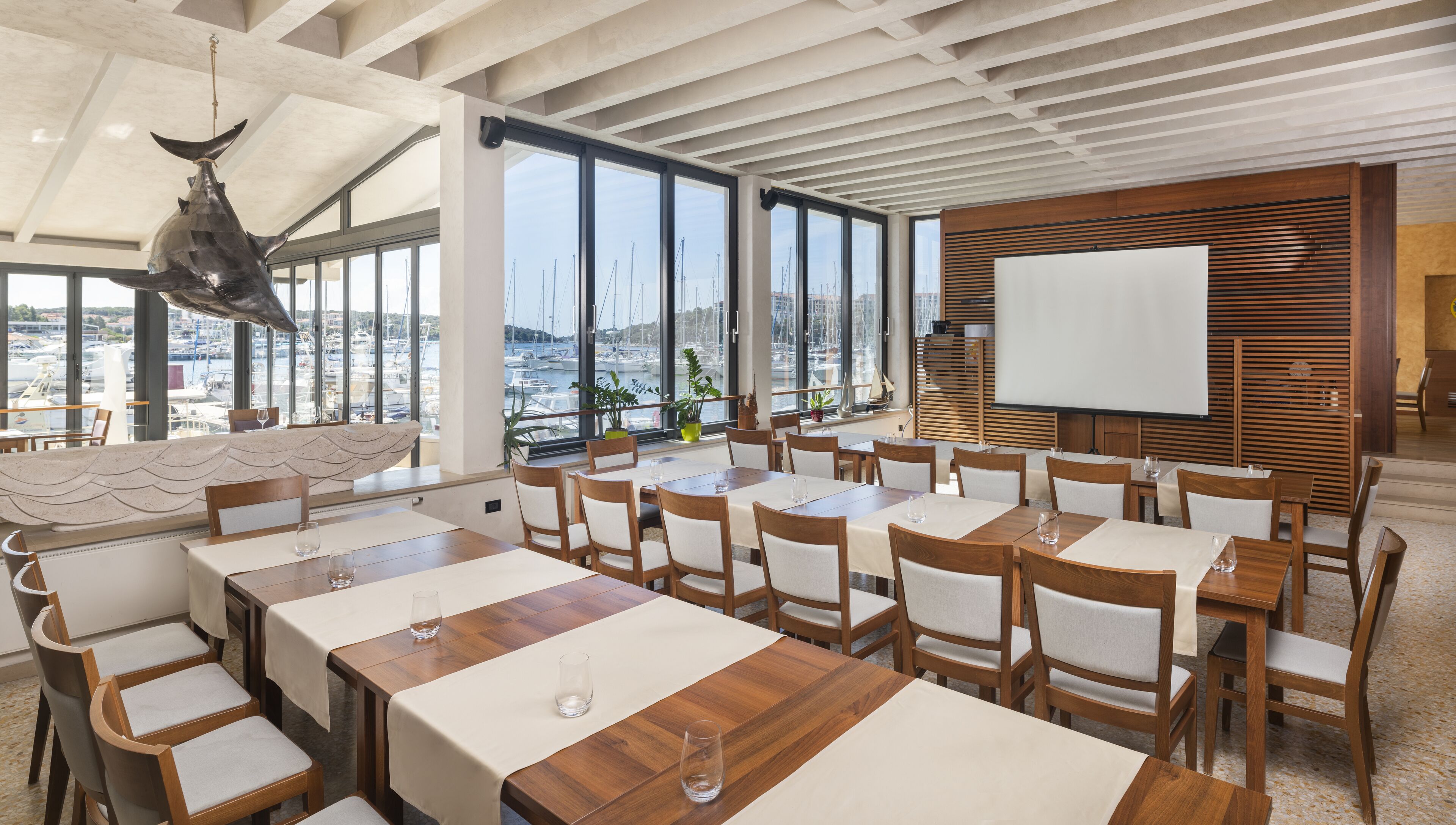 A conference room with large windows overlooking a marina, wooden tables and chairs, and a whiteboard on the wall.
