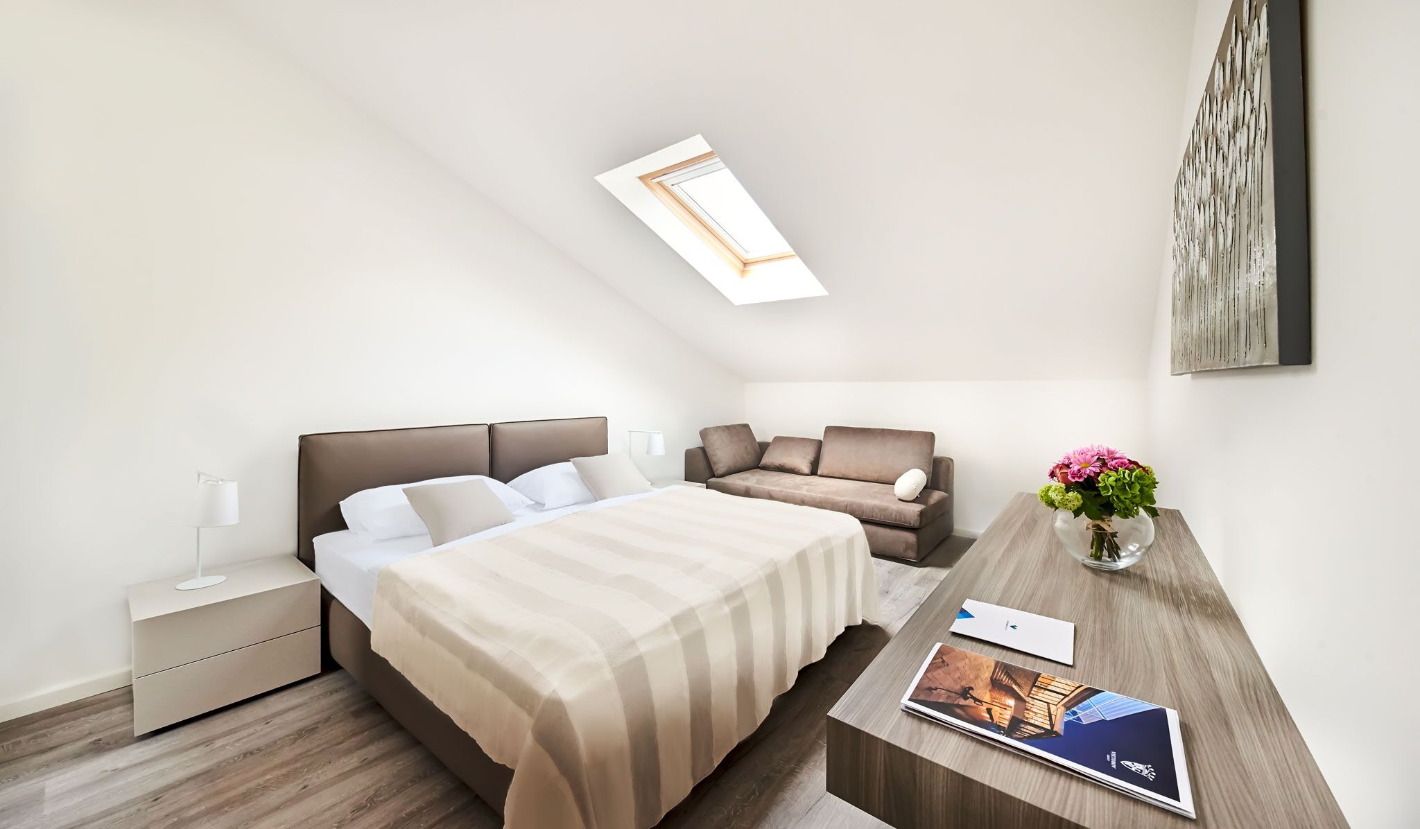 A modern hotel room with a bed, a sofa, a desk with a vase of flowers, and a skylight.