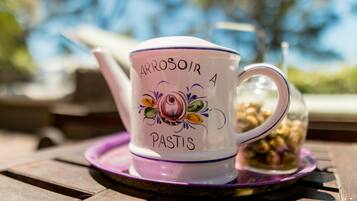 Une tasse en céramique blanche, ornée de motifs floraux, sur laquelle on peut lire les mots « ARROSOIR PASTIS ». Elle est posée sur une table avec un dessous de verre violet.