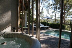 An indoor hot tub area with a wooden deck, a pool, lounge chairs, and a view of trees and hills.