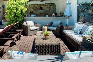 A sunlit outdoor seating area with wicker furniture and cushions, a small potted plant, and a stone wall backdrop.