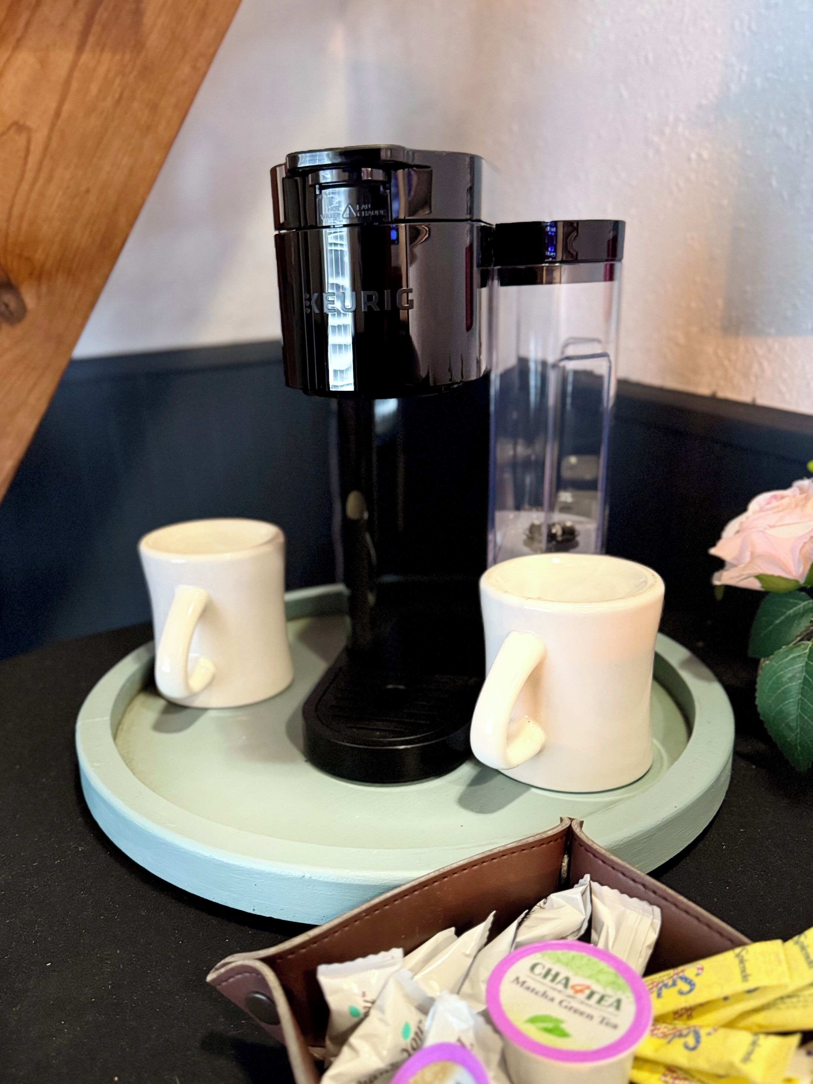 A coffee maker with two white mugs on a teal coaster, a brown leather box with tea packets, and a pink rose in the background.