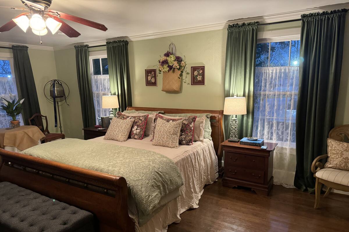 A bedroom with a wooden bed, green curtains, a ceiling fan, a bedside table with a lamp, and a window with a view.