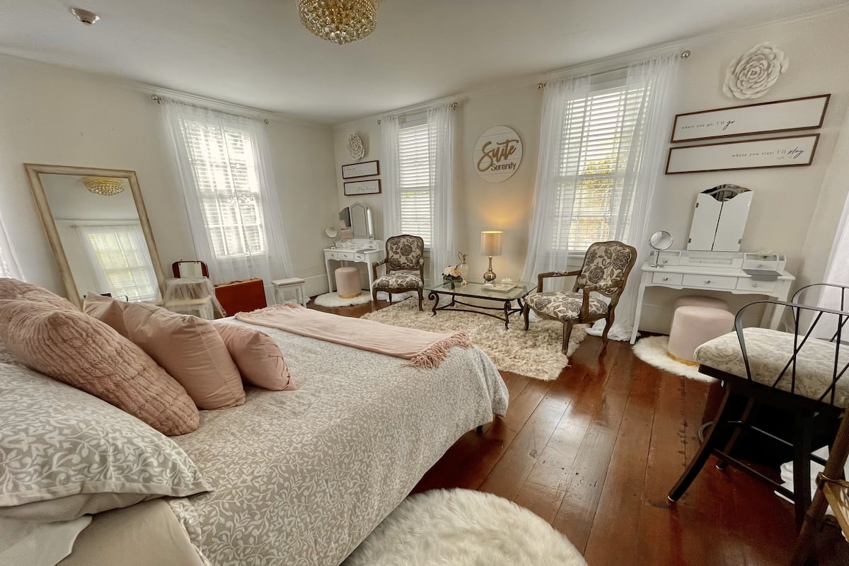 A bedroom with a large bed, a vanity with a mirror, a chair, a small table, and a dresser.