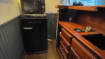 Una cocina compacta con microondas integrado, un refrigerador pequeño, armarios de madera, un lavamanos y una lámpara decorativa.