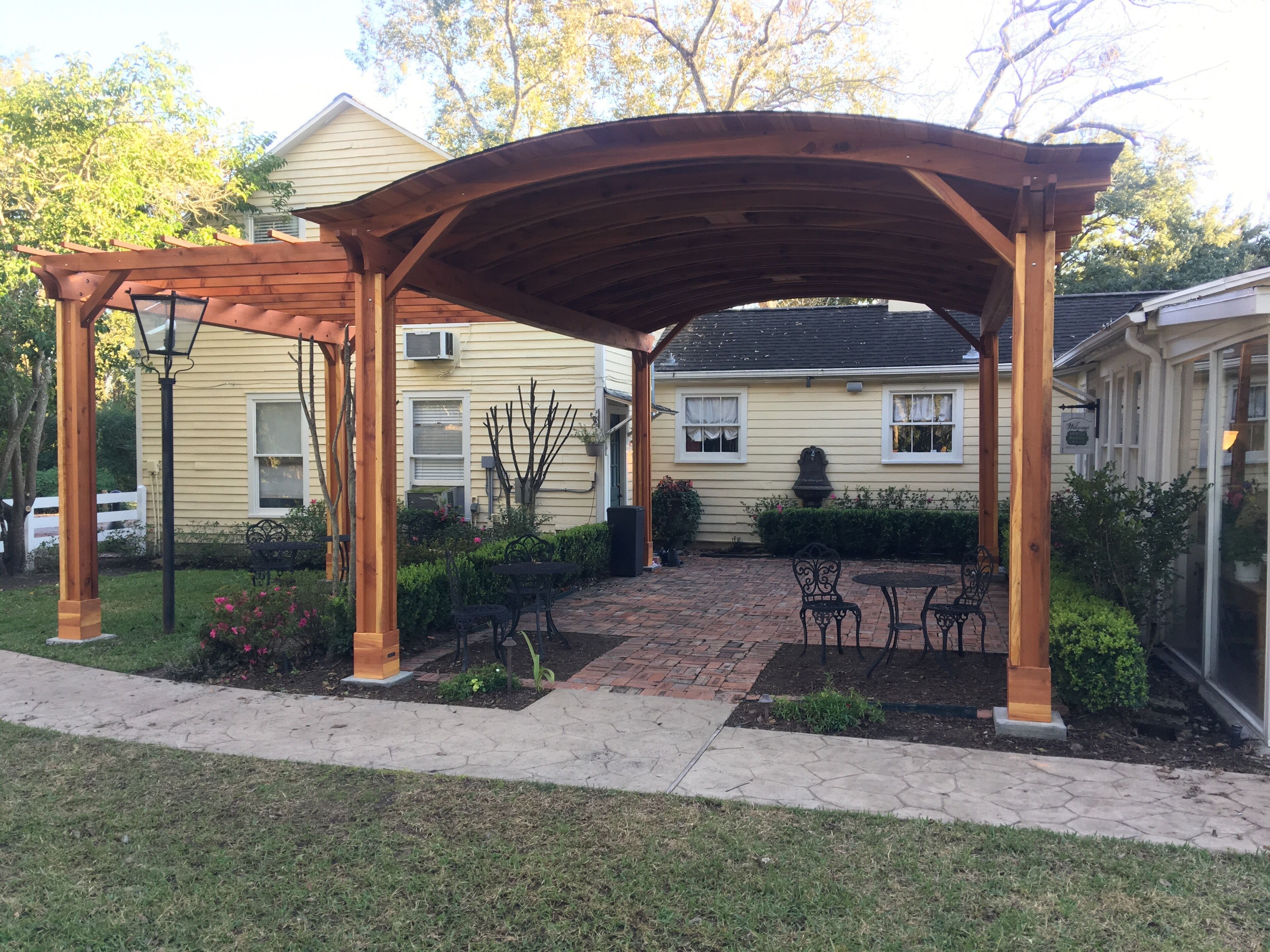 Ein überdachter Patio-Bereich mit einem Pflasterweg, einer Holz-Pergola und Gartenmöbeln.