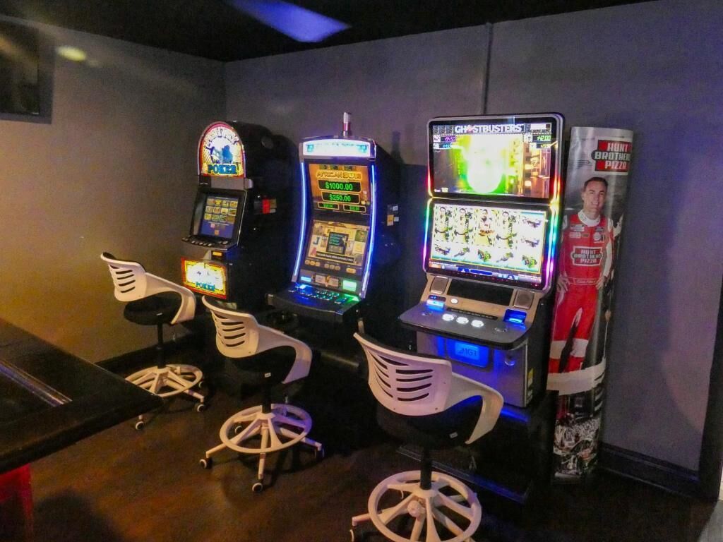 Une rangée de bornes d’arcade avec des chaises devant, une affiche accrochée au mur et un comptoir avec un menu.