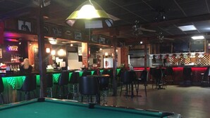 Una mesa de billar con asientos de bar alrededor y una zona de bar con un letrero de neón.