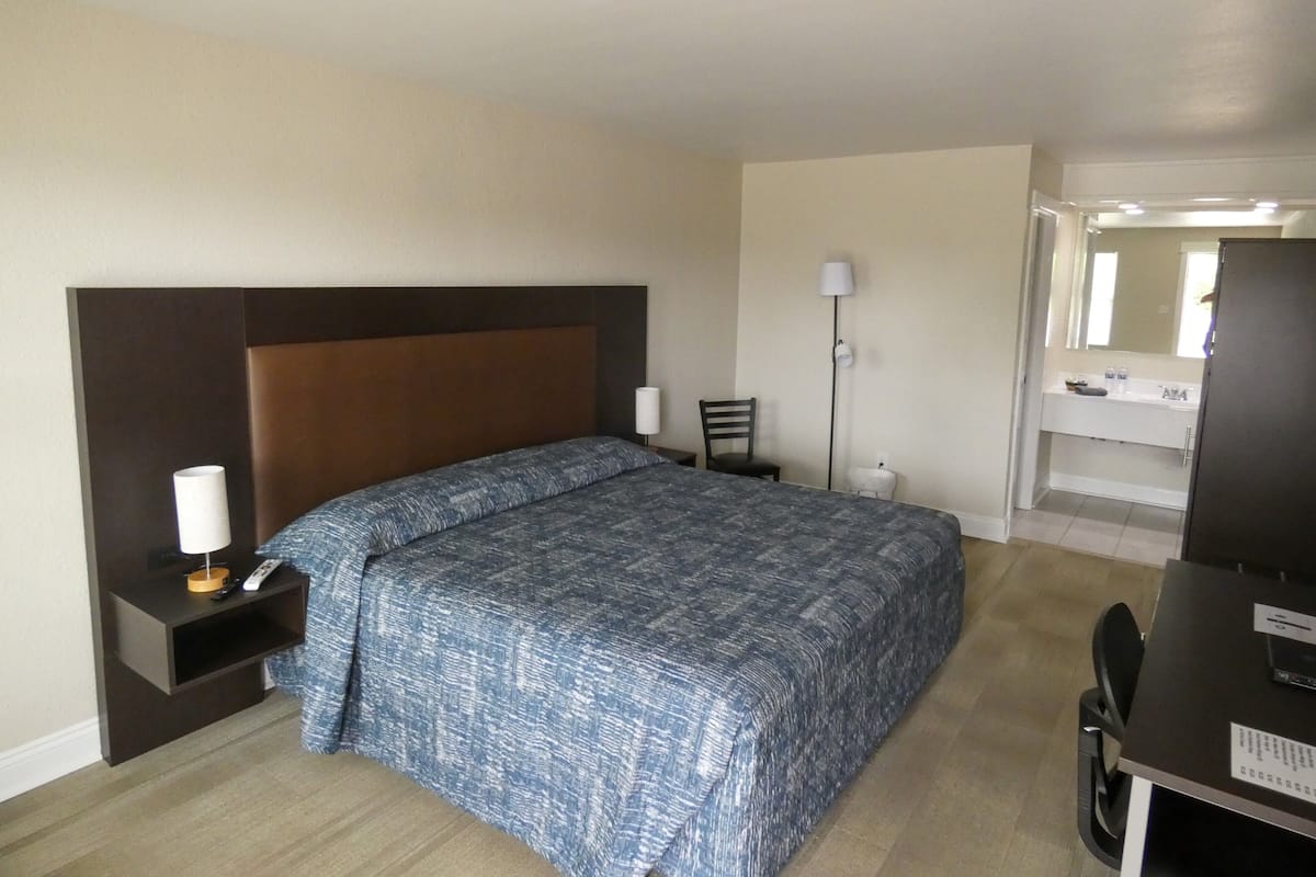 Quarto de hotel com cama, mesa de cabeceira, luminária, cadeira, escrivaninha e espelho.