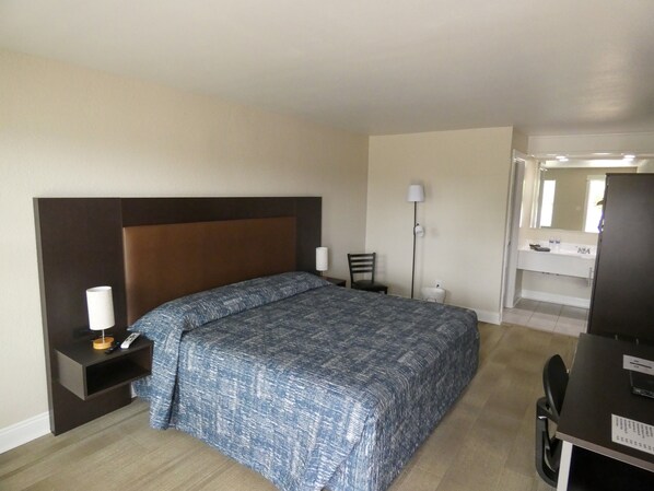Una habitación de hotel con cama, mesita de noche, lámpara, silla, escritorio y espejo.