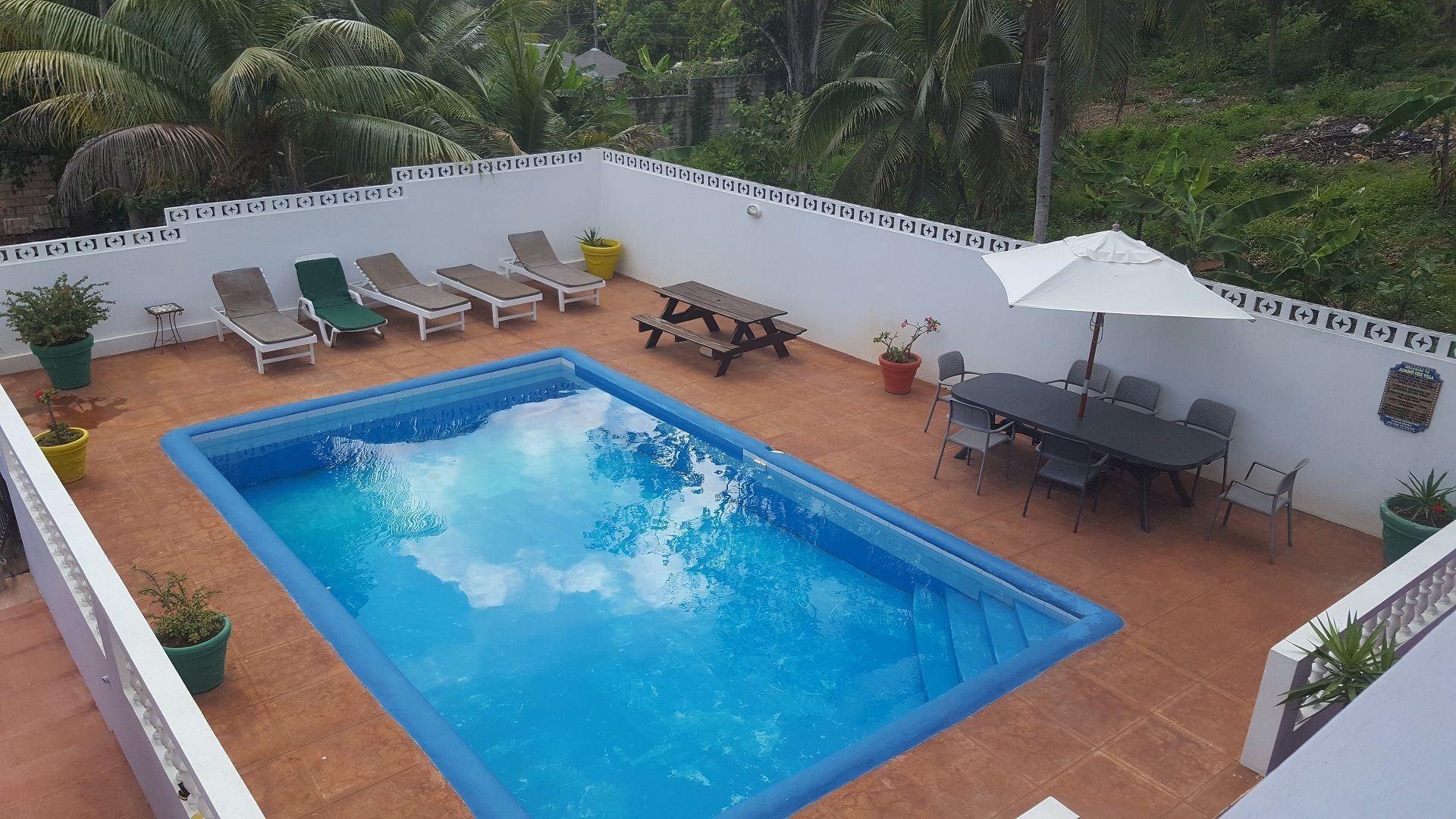 A rectangular swimming pool surrounded by lounge chairs and a table with chairs, set against a white wall with a decorative border.