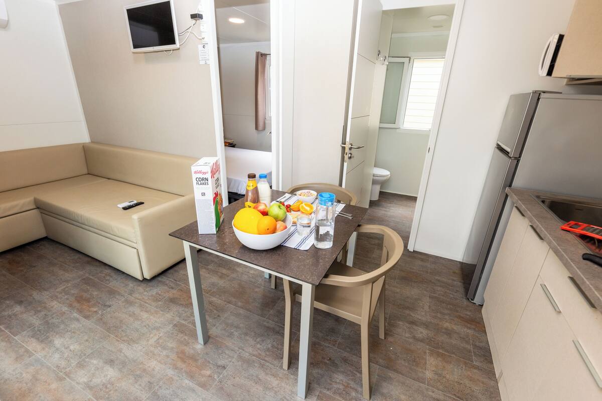 A compact living space with a kitchenette, a dining area with a table set for two, a sofa, and a small TV mounted on the wall.
