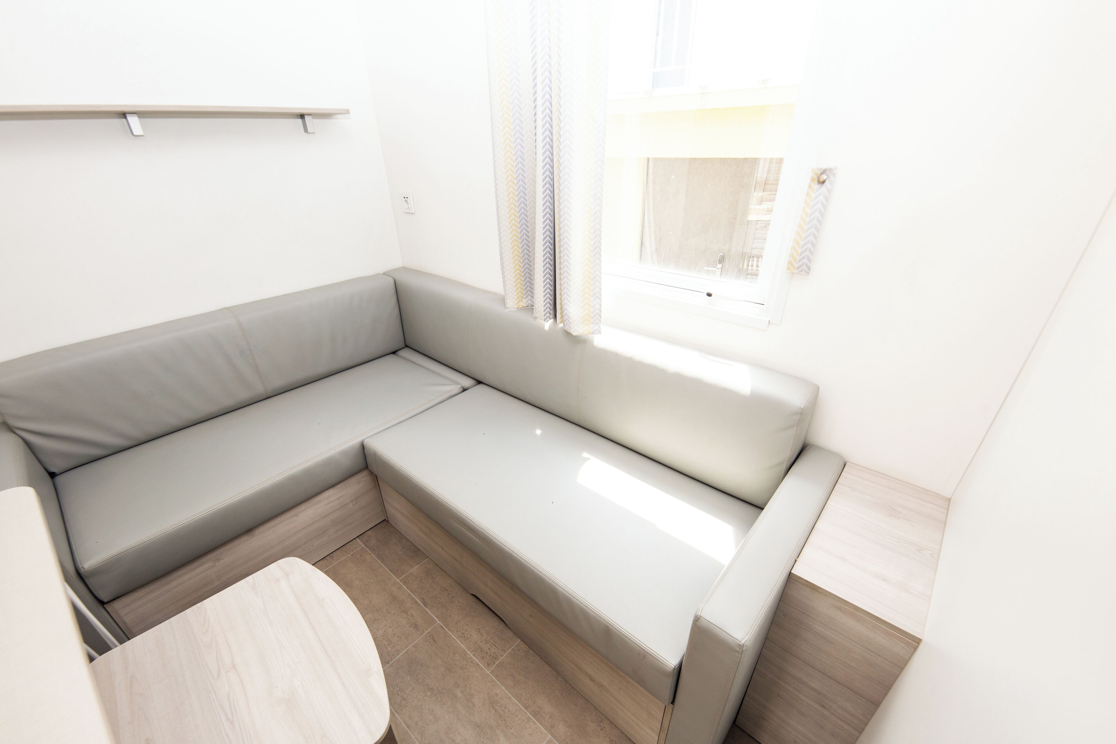 Een moderne, minimalistisch ingerichte kamer met een grijze modulaire bank, een klein houten tafeltje en een raam met gordijnen.