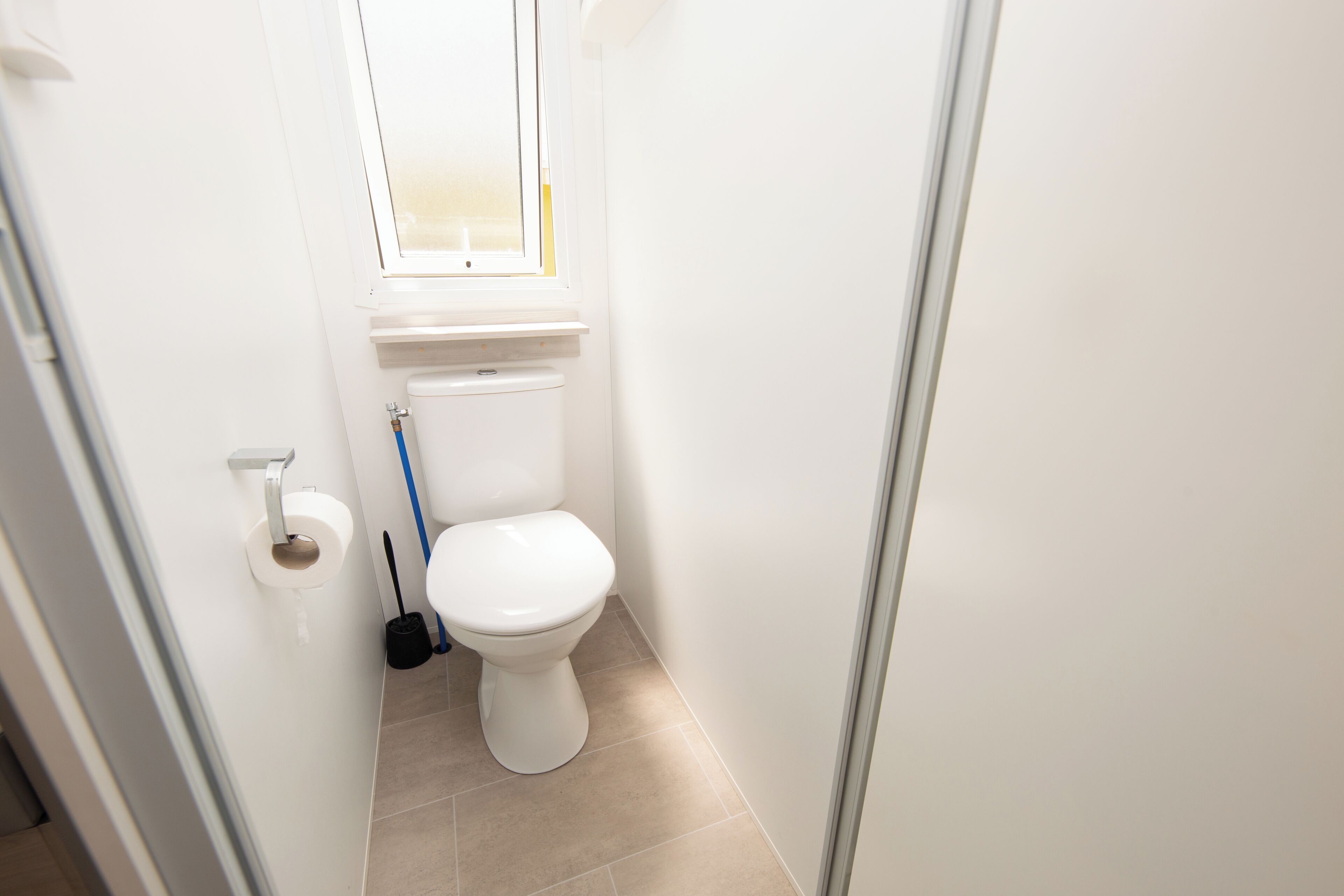 Ein kleines, sauberes Badezimmer mit Toilette, einer Toilettenpapierrolle und einem an der Wand angelehnten Besen.