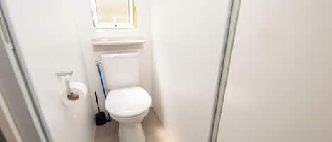 Un baño pequeño y limpio con inodoro, un rollo de papel higiénico y una escoba apoyada en la pared.