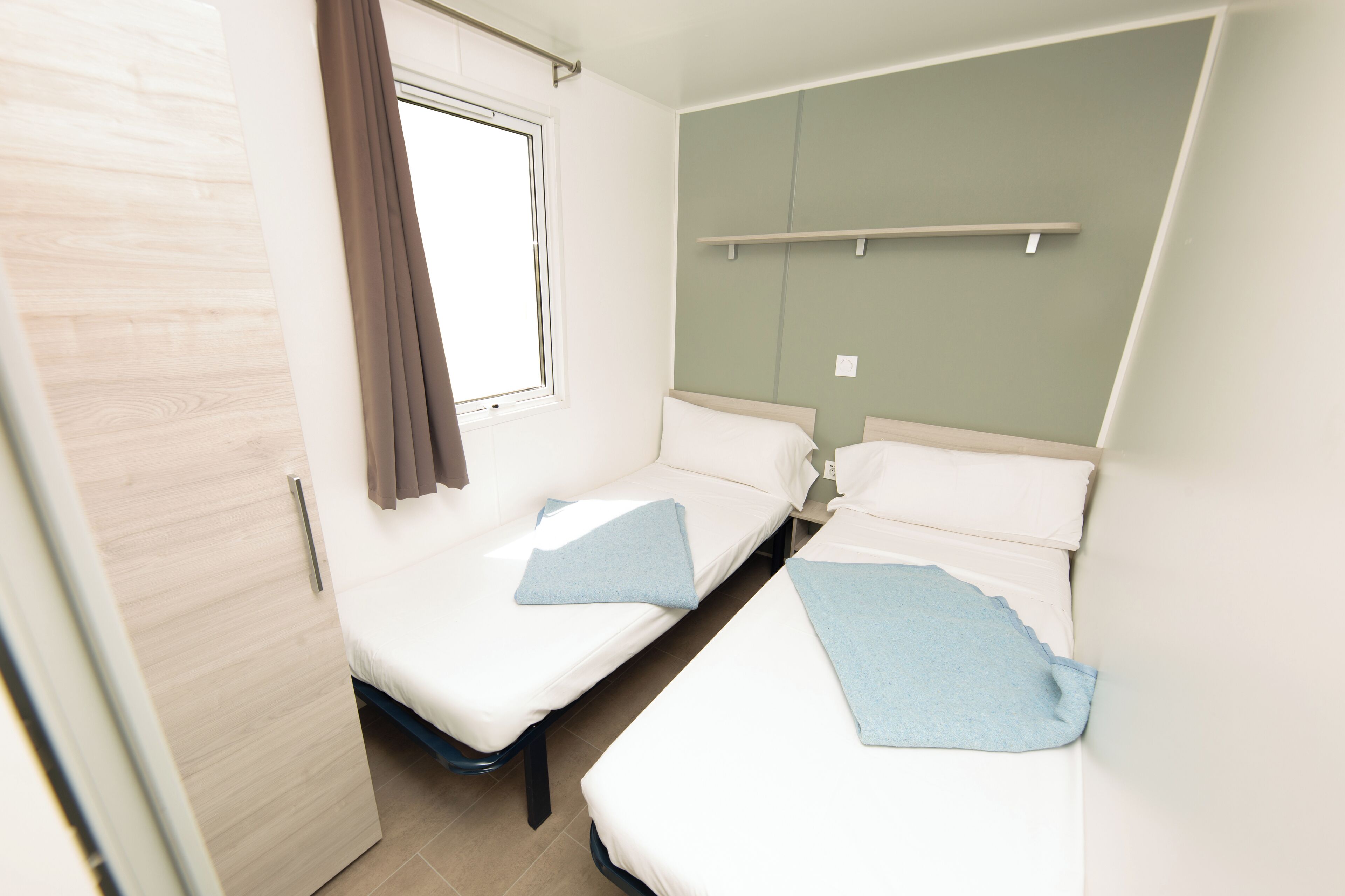 A small, clean room with two single beds, a window with curtains, a wooden door, and a light-colored wall.