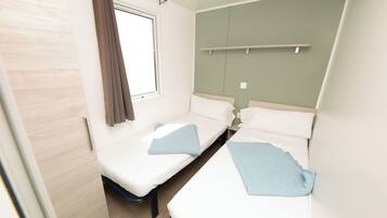 A small, clean room with two single beds, a window with curtains, a wooden door, and a light-colored wall.