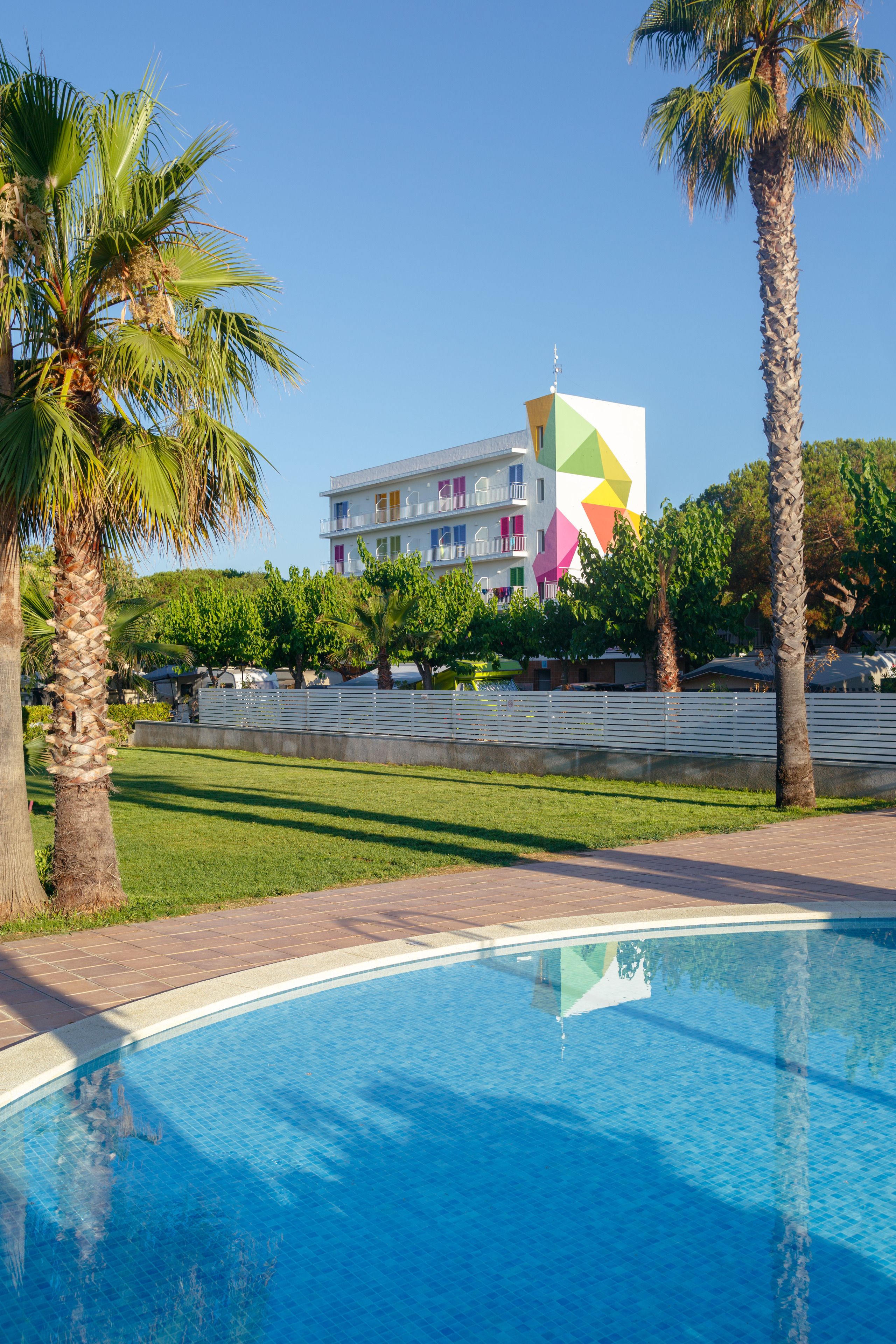 A swimming pool with palm trees, a grassy area, and a colorful building in the background.