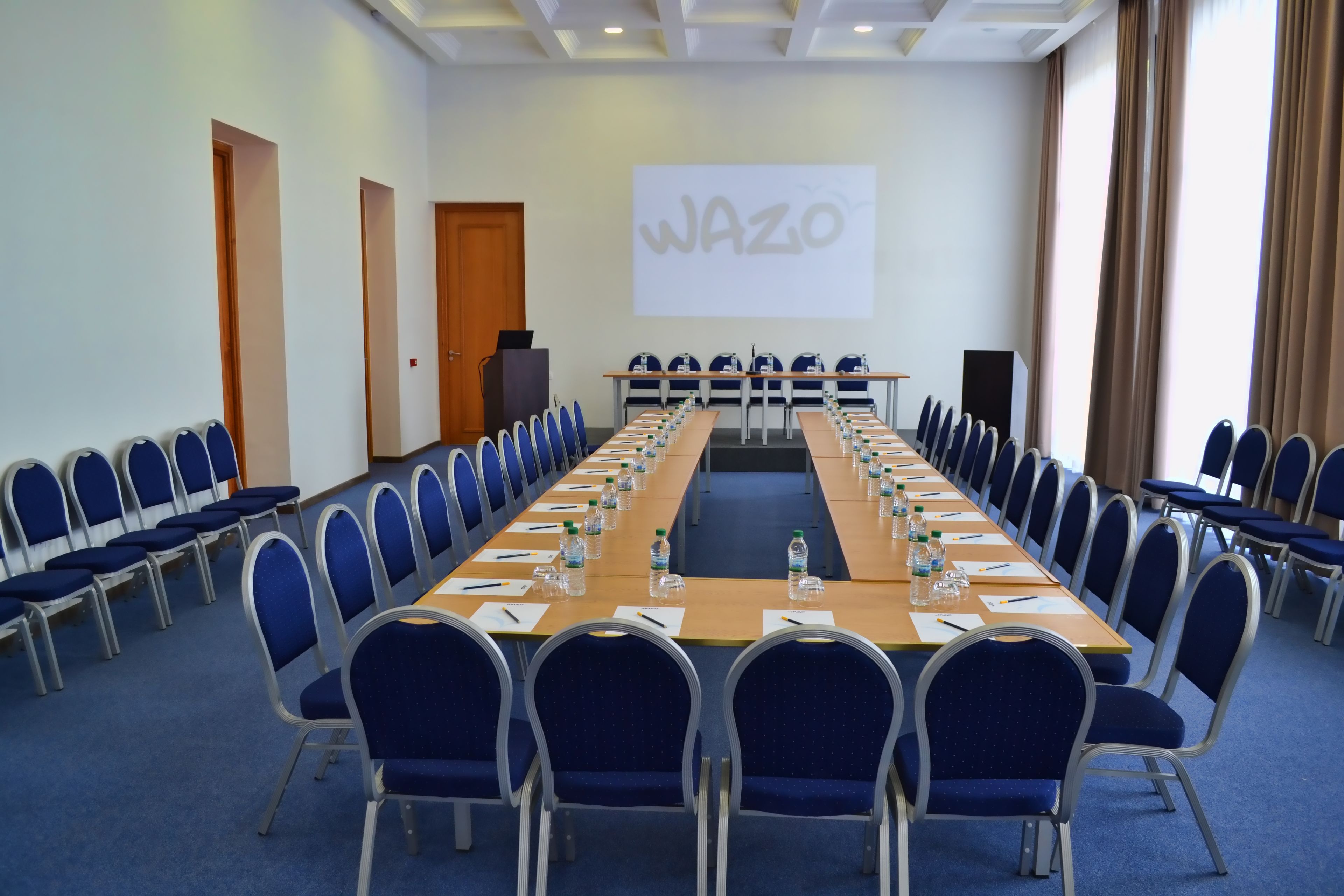 Un salón de conferencias con una larga mesa, sillas azules y una pantalla de proyector mostrando 'WAZO'.