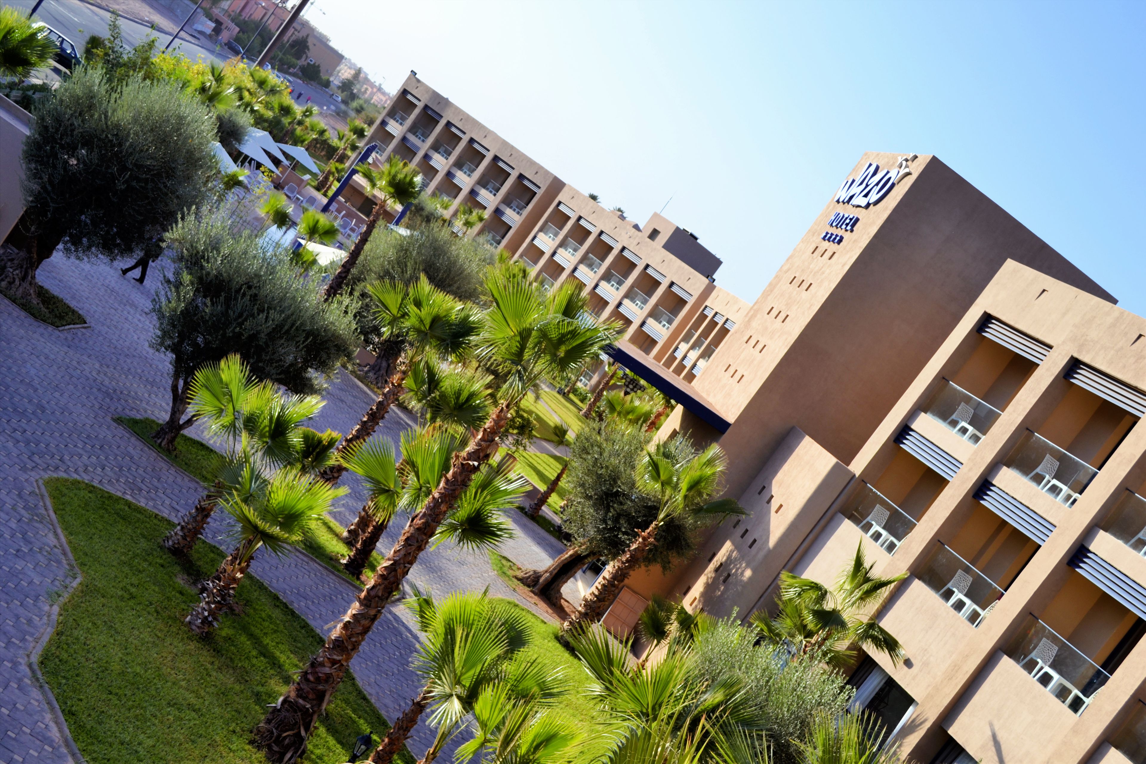 A modern hotel with a flat roof, multiple balconies, and a central courtyard with palm trees and a fountain.