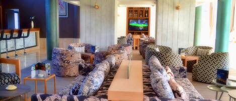 Una sala de estar moderna con sofás estampados, una mesa de madera larga y un televisor al fondo.
