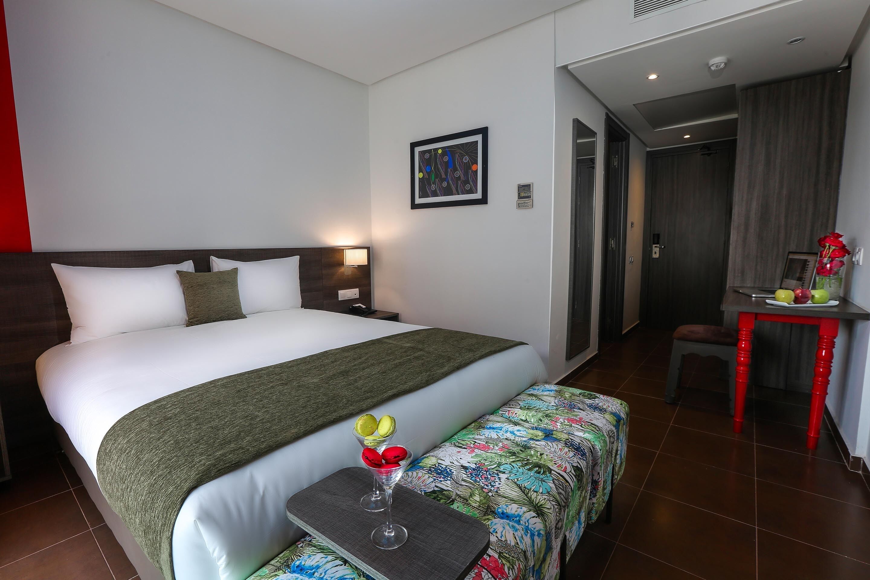 A hotel room with a bed, a bedside table with a drink, a bench, a desk with a chair, and a door leading to another room.