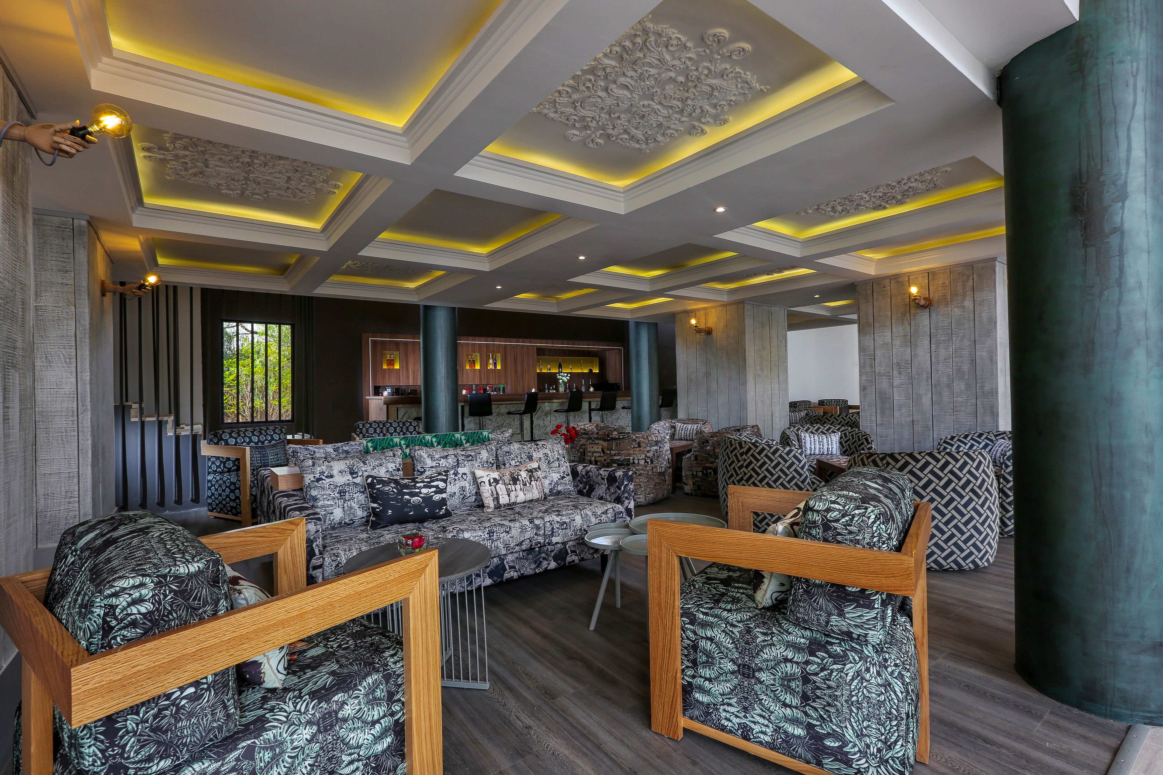 Un vaste espace salon agrémenté de canapés à motifs et de chaises en bois, un bar avec des tabourets et un plafond décoratif doté d’un éclairage encastré.