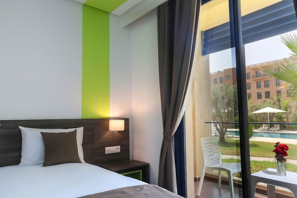 A hotel room with a bed, a bedside table with a lamp, a window with curtains, and a view of a pool and buildings outside.