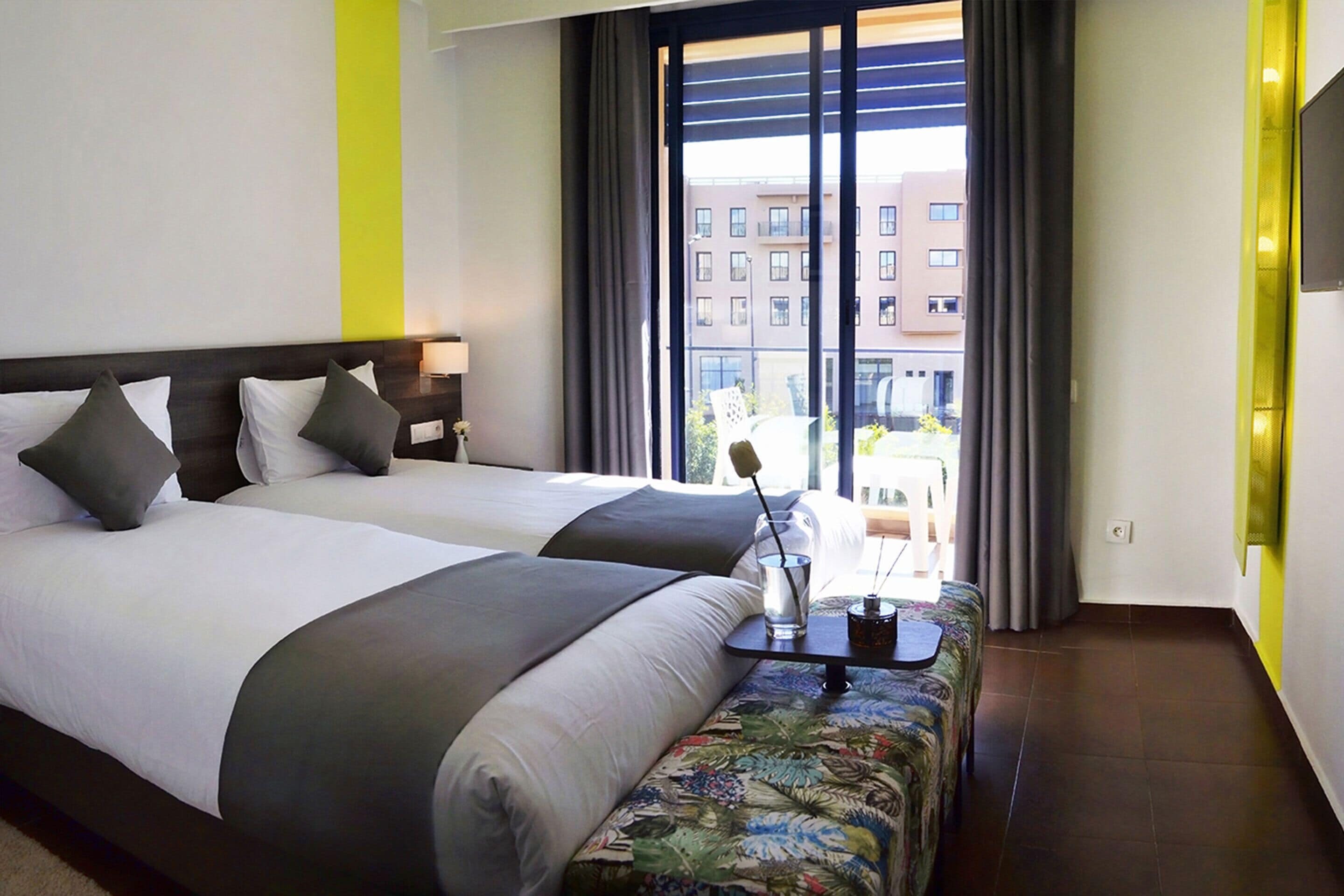Ein Hotelzimmer mit zwei Betten, einem kleinen Tisch mit einem Getränk und einem Notizblock, und Blick auf Gebäude vor dem Fenster.