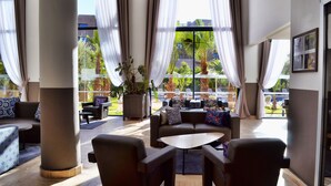 Un vaste espace salon doté d’une grande fenêtre offrant une vue sur des palmiers et un immeuble. On y trouve plusieurs canapés et fauteuils disposés autour d’une table basse centrale.