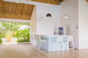 A bar area with a tiled backsplash, white bar stools, and pendant lighting.