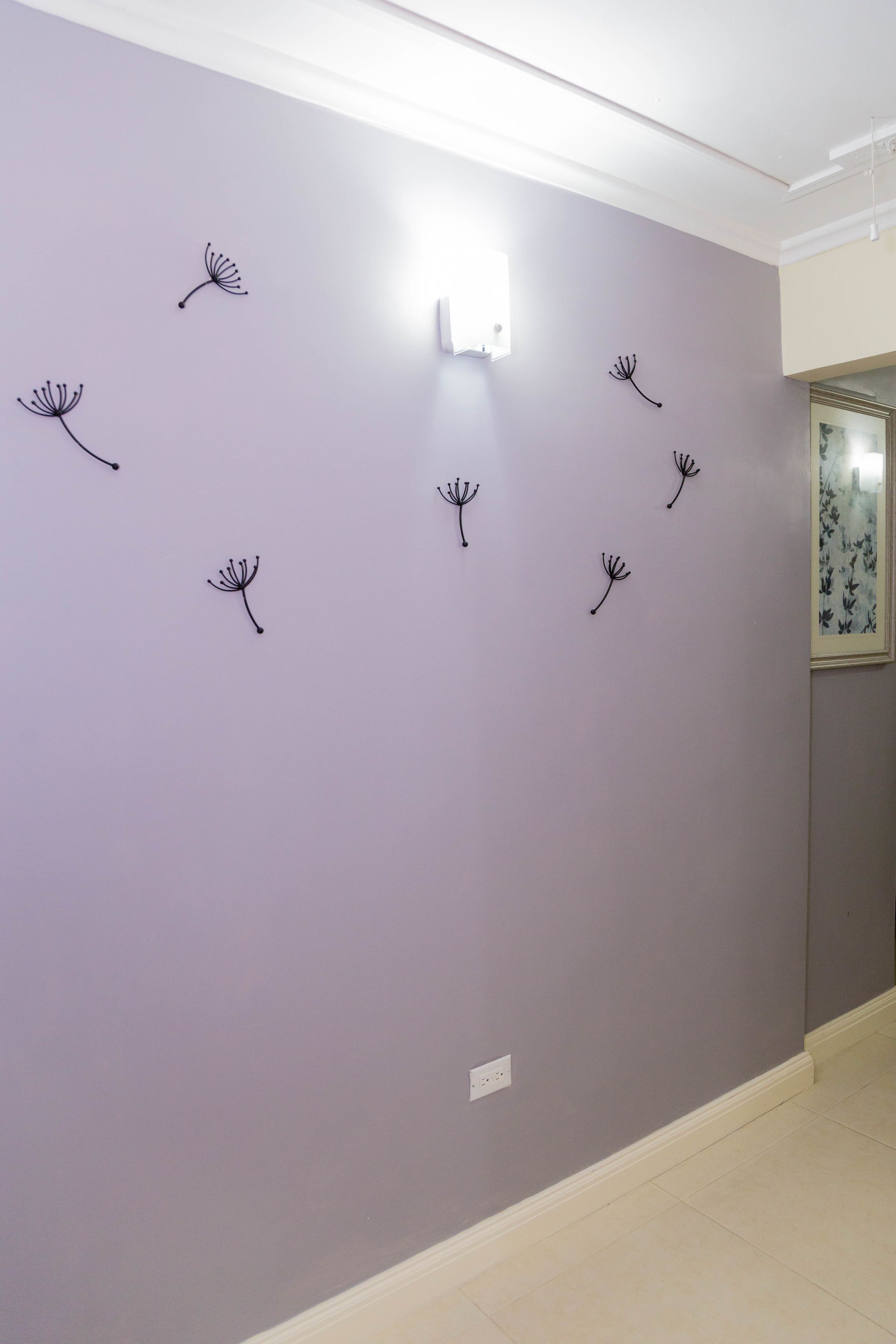 Un couloir avec un mur de couleur violet pâle, un plafond blanc et un luminaire. On y trouve six éléments décoratifs noirs fixés au mur, qui ressemblent à des graines de pissenlit.