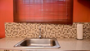 Una cocina con un lavamanos de acero inoxidable, un salpicadero de azulejos y un ventanal con persianas.