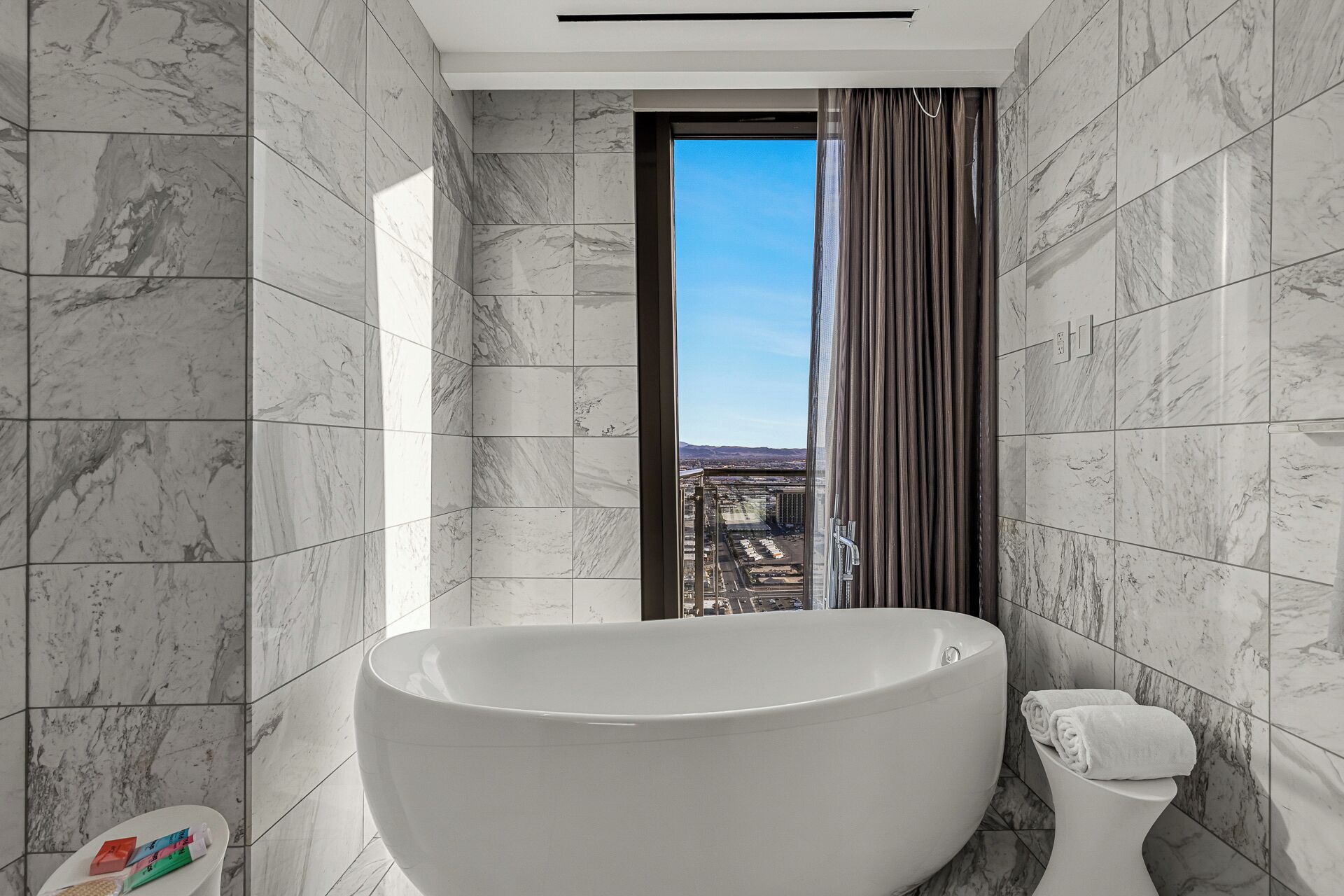 Een moderne badkamer met een vrijstaand bad, een toilet en uitzicht op een stadsgezicht door een raam.
