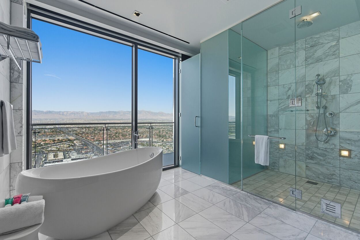 A modern bathroom with a freestanding bathtub, a glass-enclosed shower, and a city view through large windows.