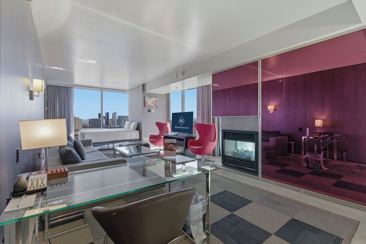 Et moderne hotelværelse med et glas skrivebord, en sofa, en pejs og udsigt over byen.
