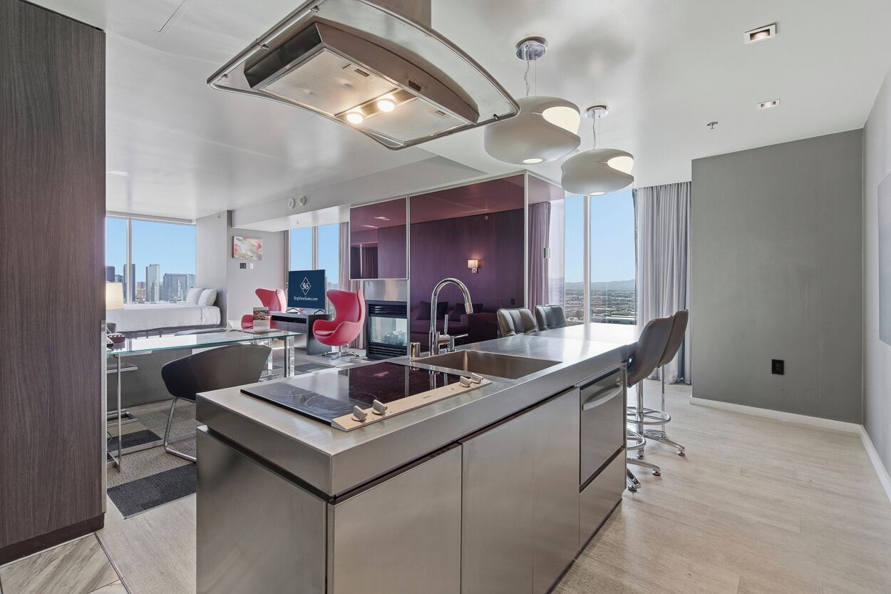 A modern kitchen with a central island, a dining area with a red chair, and a view of the cityscape.