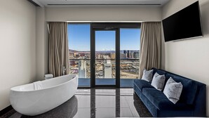 Habitación de hotel moderna con un amplio ventanal corredero que da a vistas de la ciudad, una bañera blanca, un sofá azul oscuro y un televisor de pantalla plana montado en la pared.