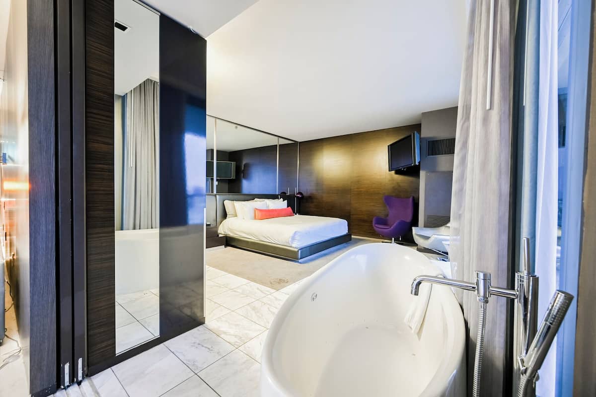 Une chambre d’hôtel moderne avec une grande baignoire, un lit avec du linge de maison blanc, un fauteuil violet et un grand miroir.