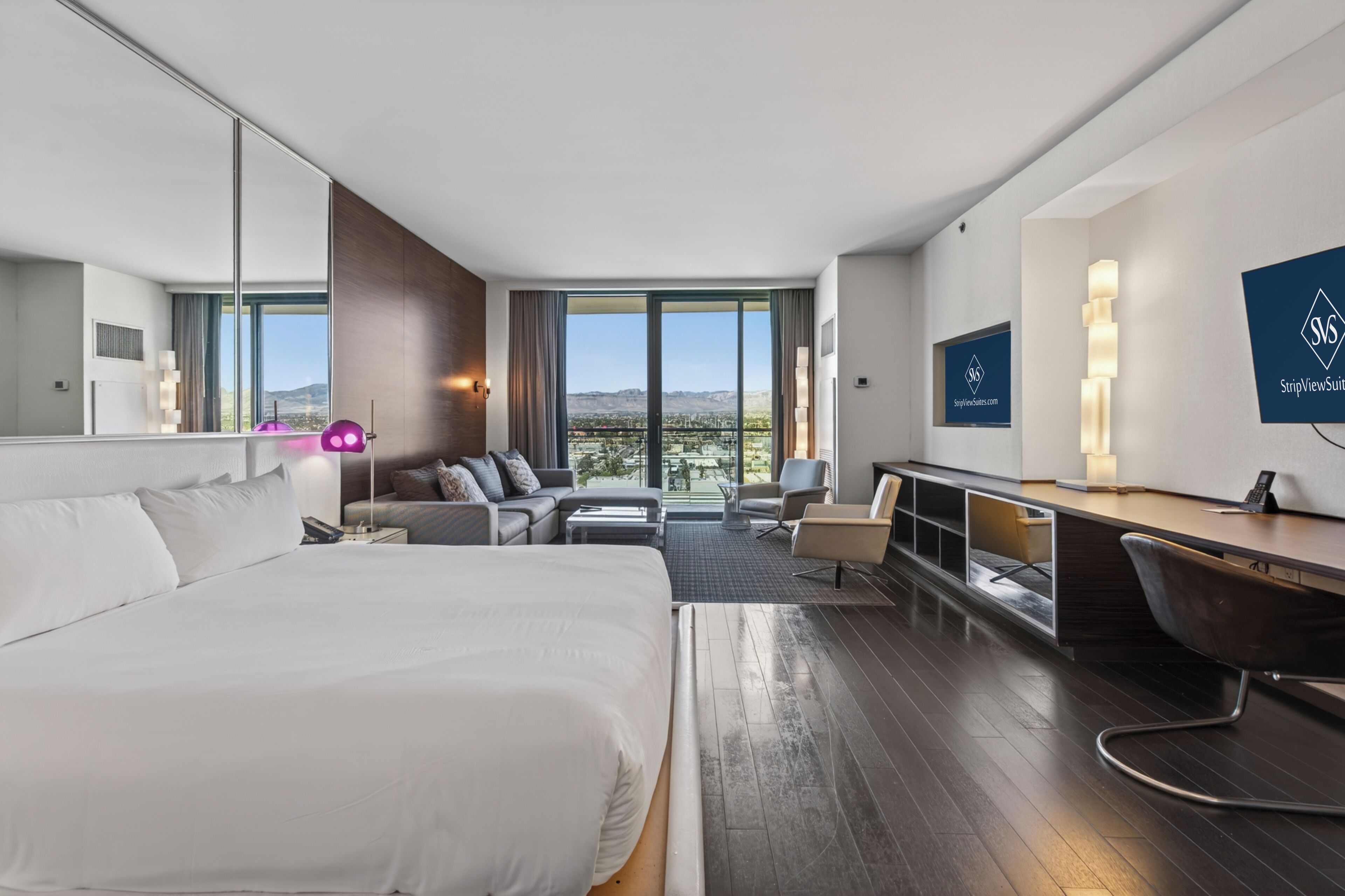 A modern hotel room with a large bed, a desk, a chair, a sofa, and a view of the city.