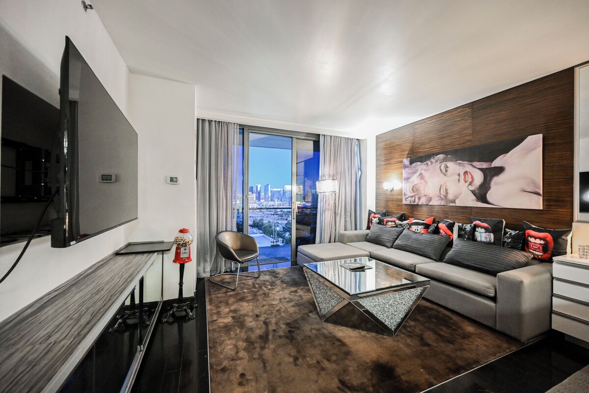 A modern hotel room with a large flat-screen TV, a sofa with decorative pillows, a glass coffee table, and a city view through large windows.